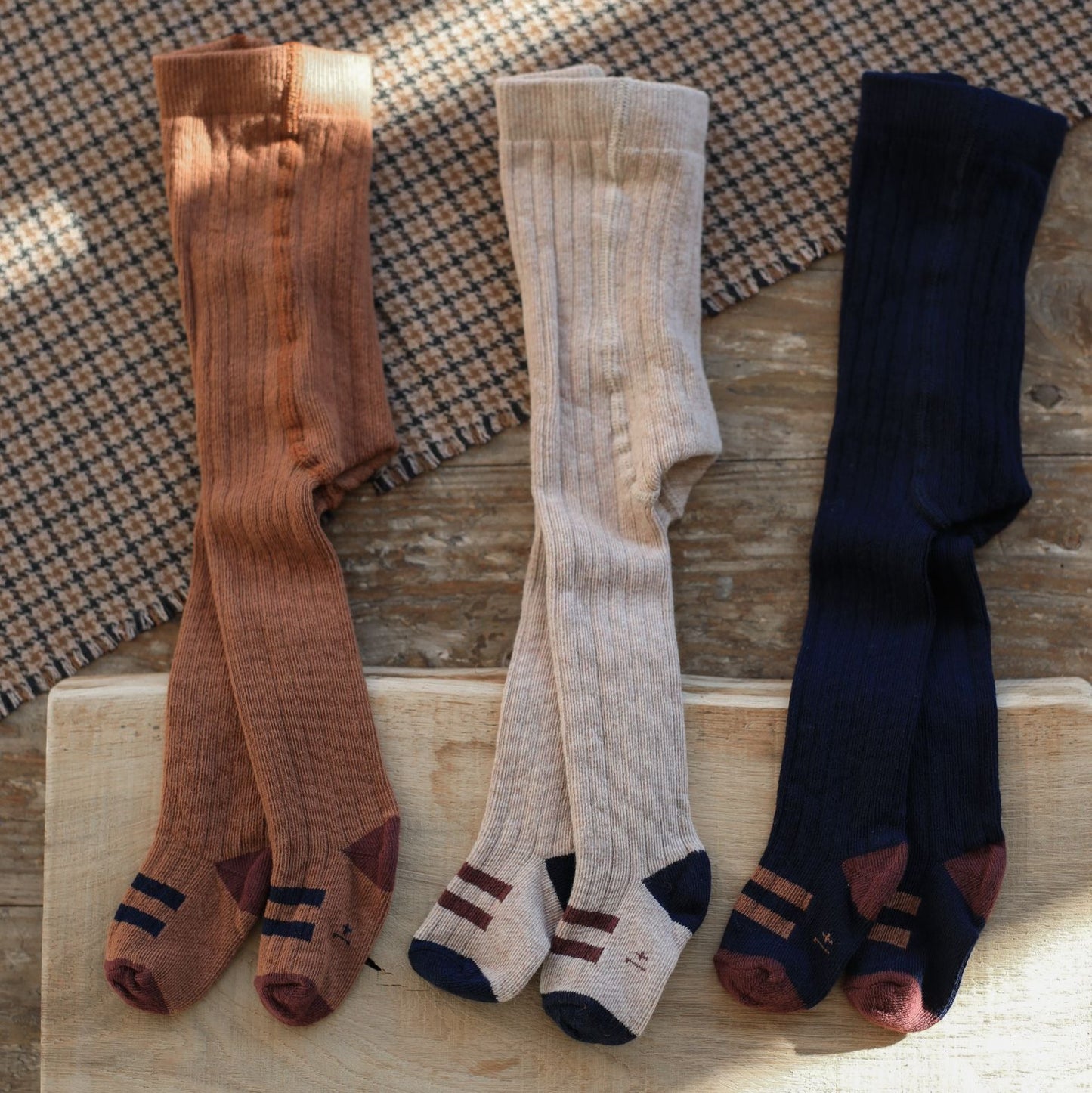 Three pairs of socks in brown, beige, and navy on a wooden surface with a checkered fabric background.