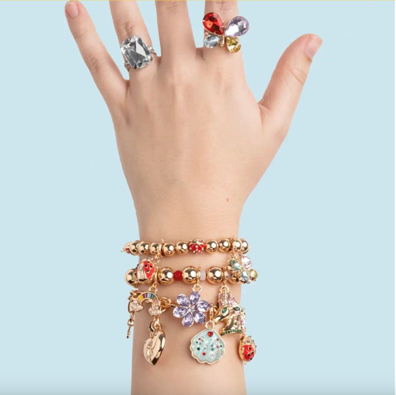 Hand wearing multiple bracelets and rings with colorful charms on a light blue background
