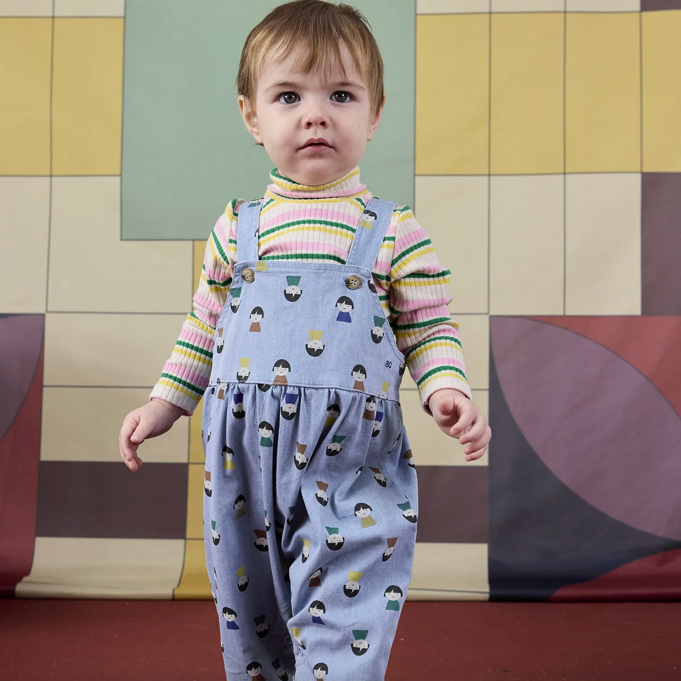 Child wearing a patterned outfit standing in front of a colorful geometric wall.
