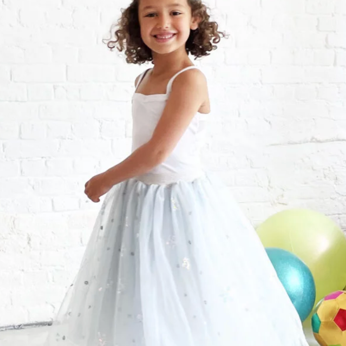 Young girl in a white dress standing next to colorful balloons against a white brick wall.