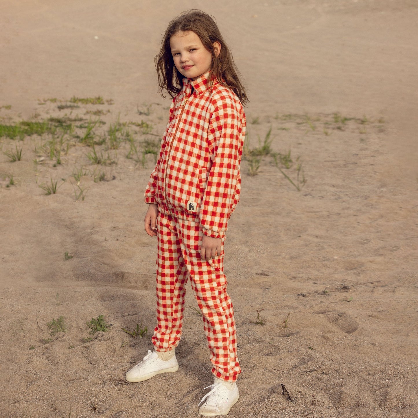 Child wearing a red and white checkered outfit standing on sandy ground.