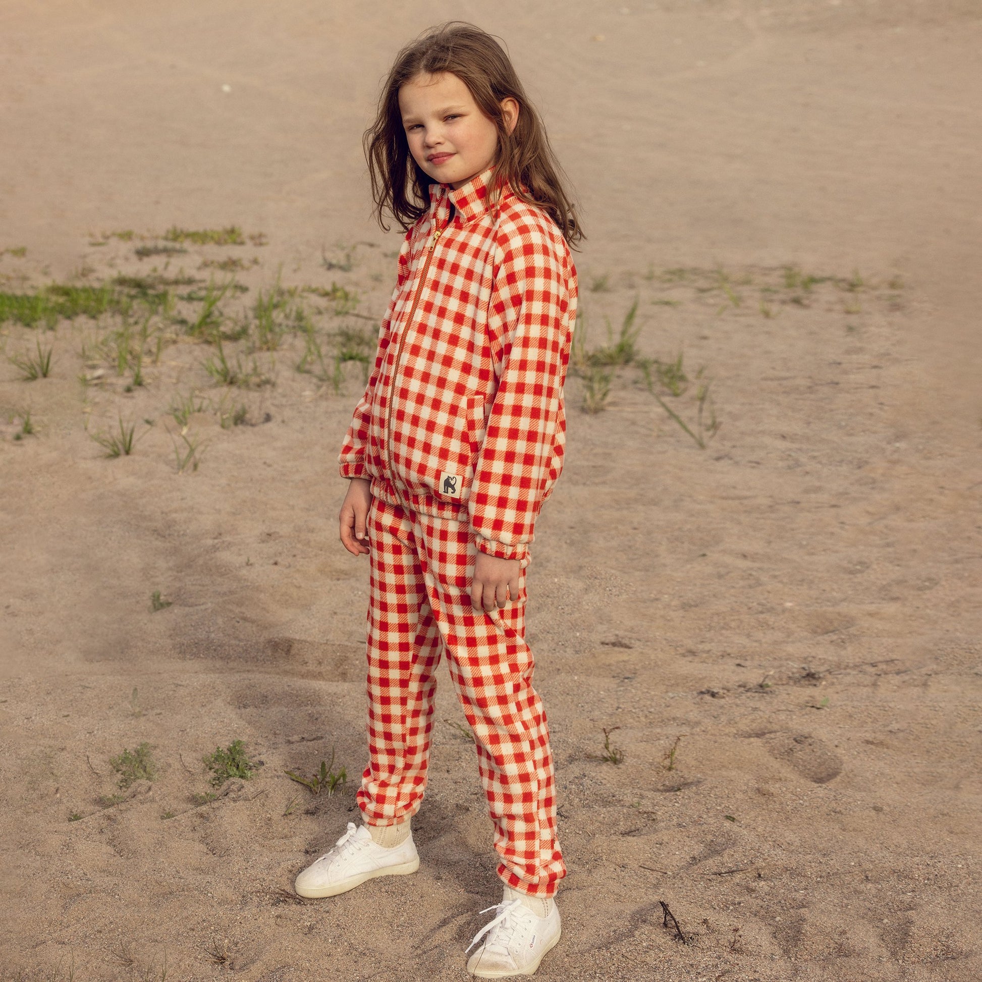 Child wearing a red and white checkered outfit standing on sandy ground.
