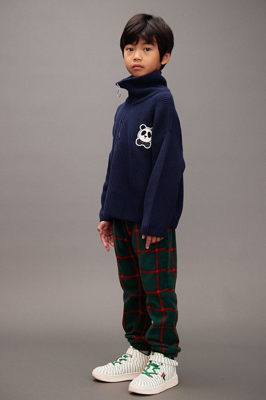 Child wearing a navy sweater with a panda design and plaid fleece pants on a green background