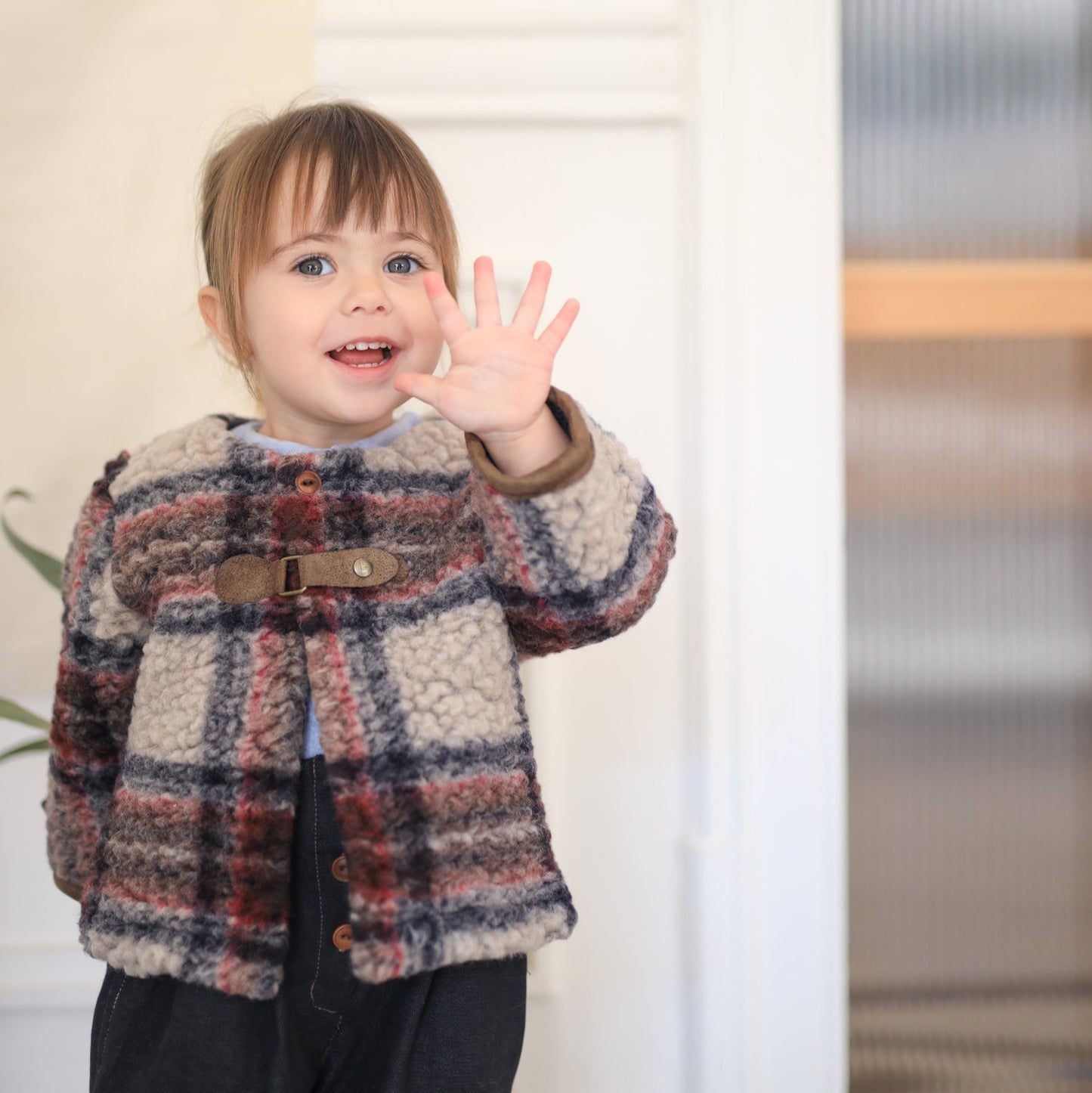 Child wearing a plaid coat waving at the camera indoors.