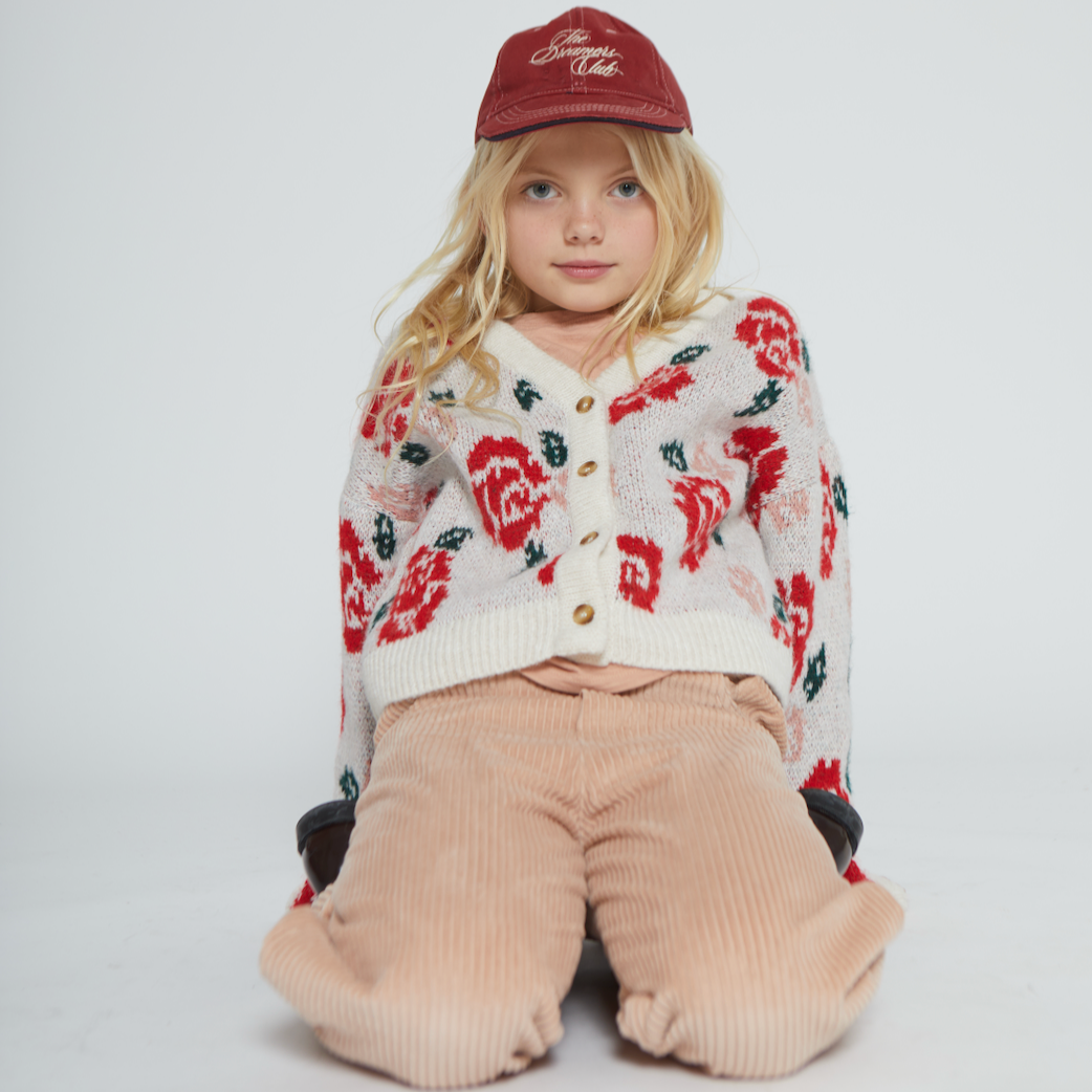 Child wearing a floral cardigan and beige pants with a red cap on a white background