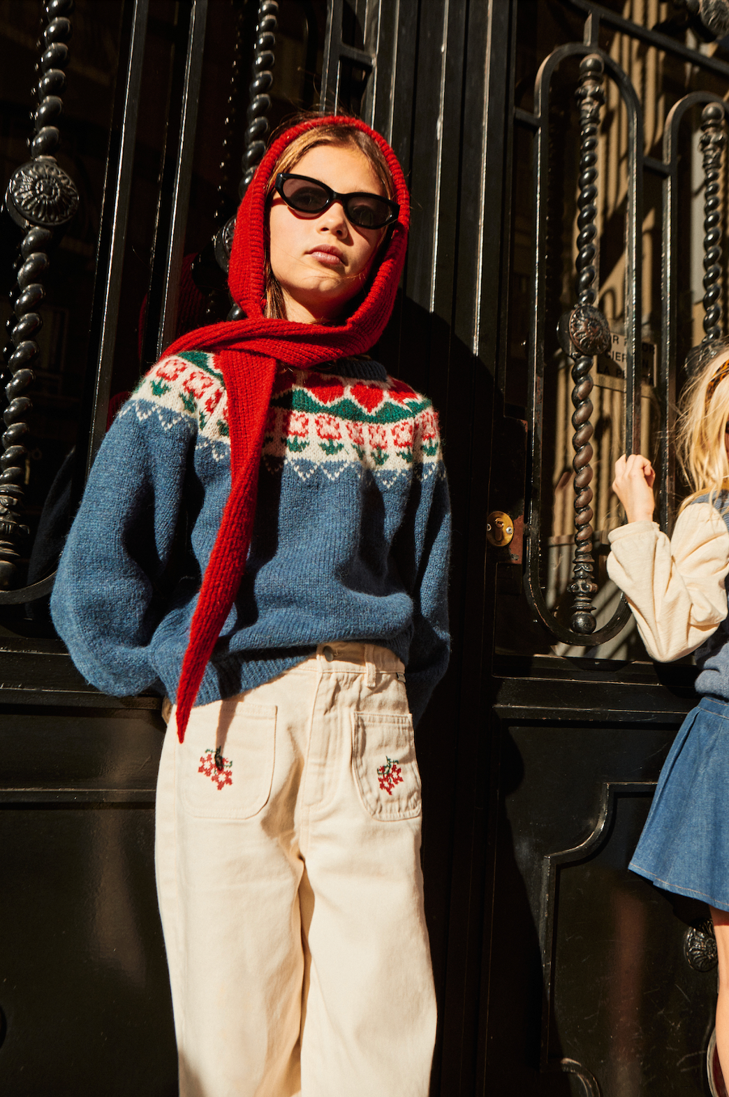 Person wearing a blue sweater with a red scarf and sunglasses, standing in front of a black metal gate.