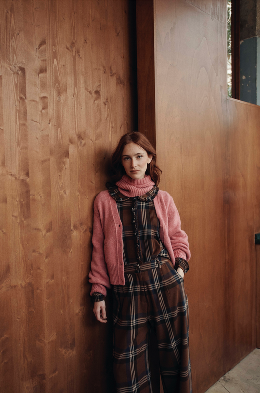Woman in a pink cardigan and plaid dress standing against a wooden wall.