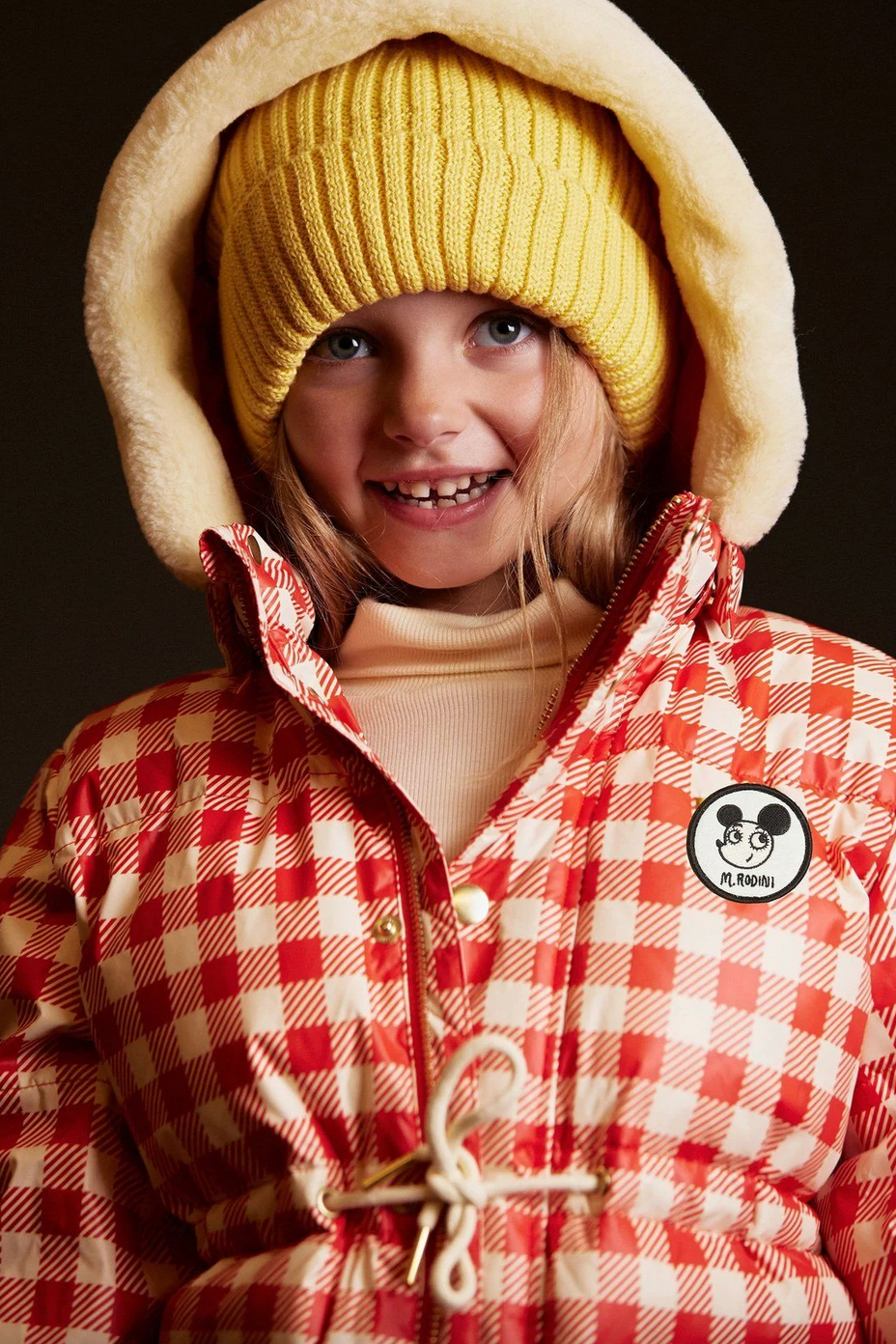 Child wearing a red checkered jacket with a hood and yellow knit hat on a dark background