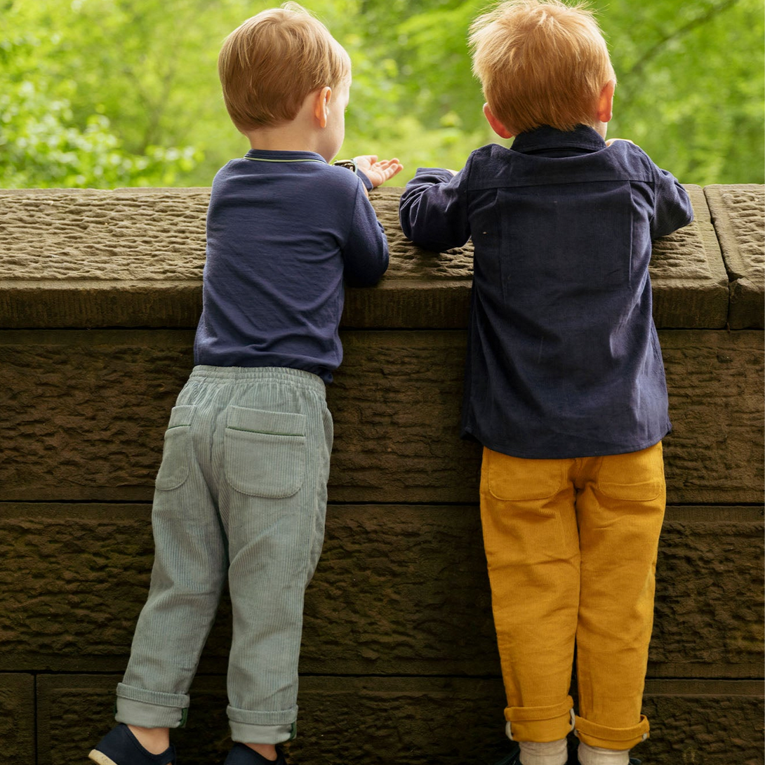 Two children standing on a wooden platform looking over a railing with greenery in the background.