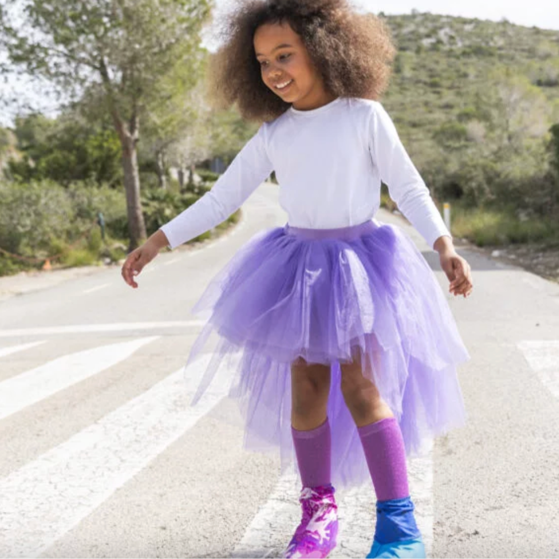 Child wearing a purple tutu skirt standing on a road with greenery in the background