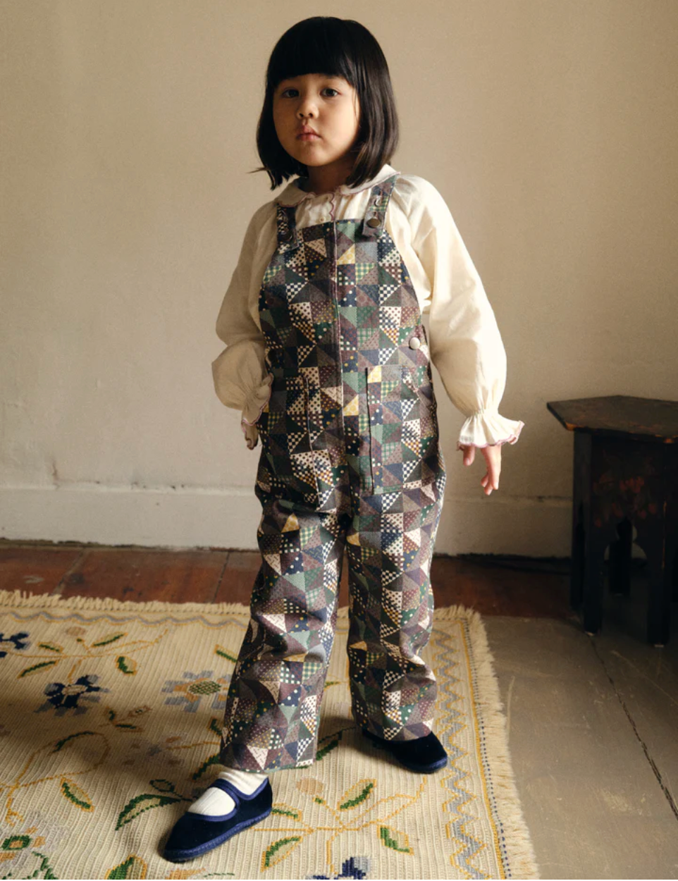 Child wearing a patterned outfit standing on a rug with a neutral background
