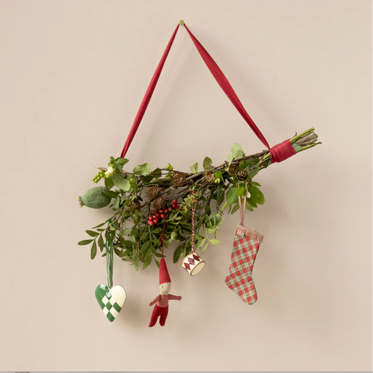 Decorative wall hanging with greenery, small figurines, and red ribbons on a beige background