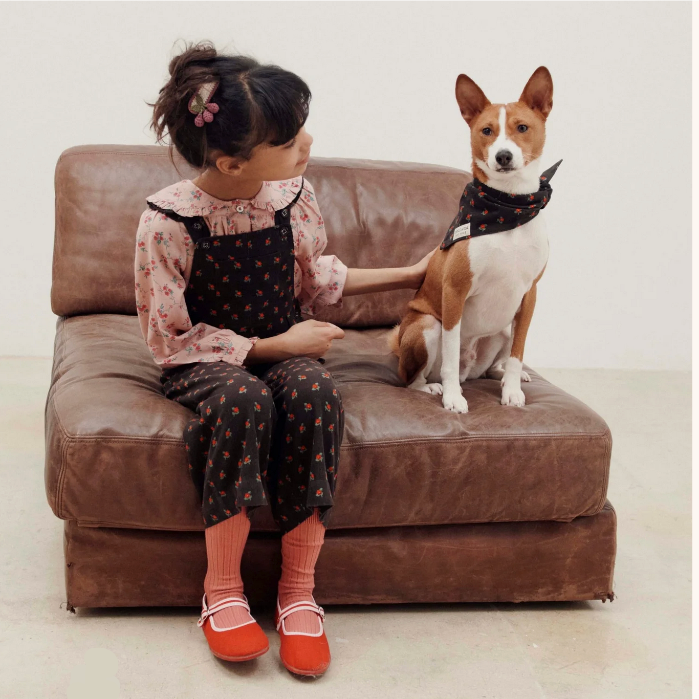 Child sitting on a brown couch with a dog wearing a bandana.