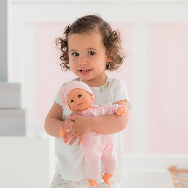 Child holding a baby doll in a room with a pink wall and white furniture.